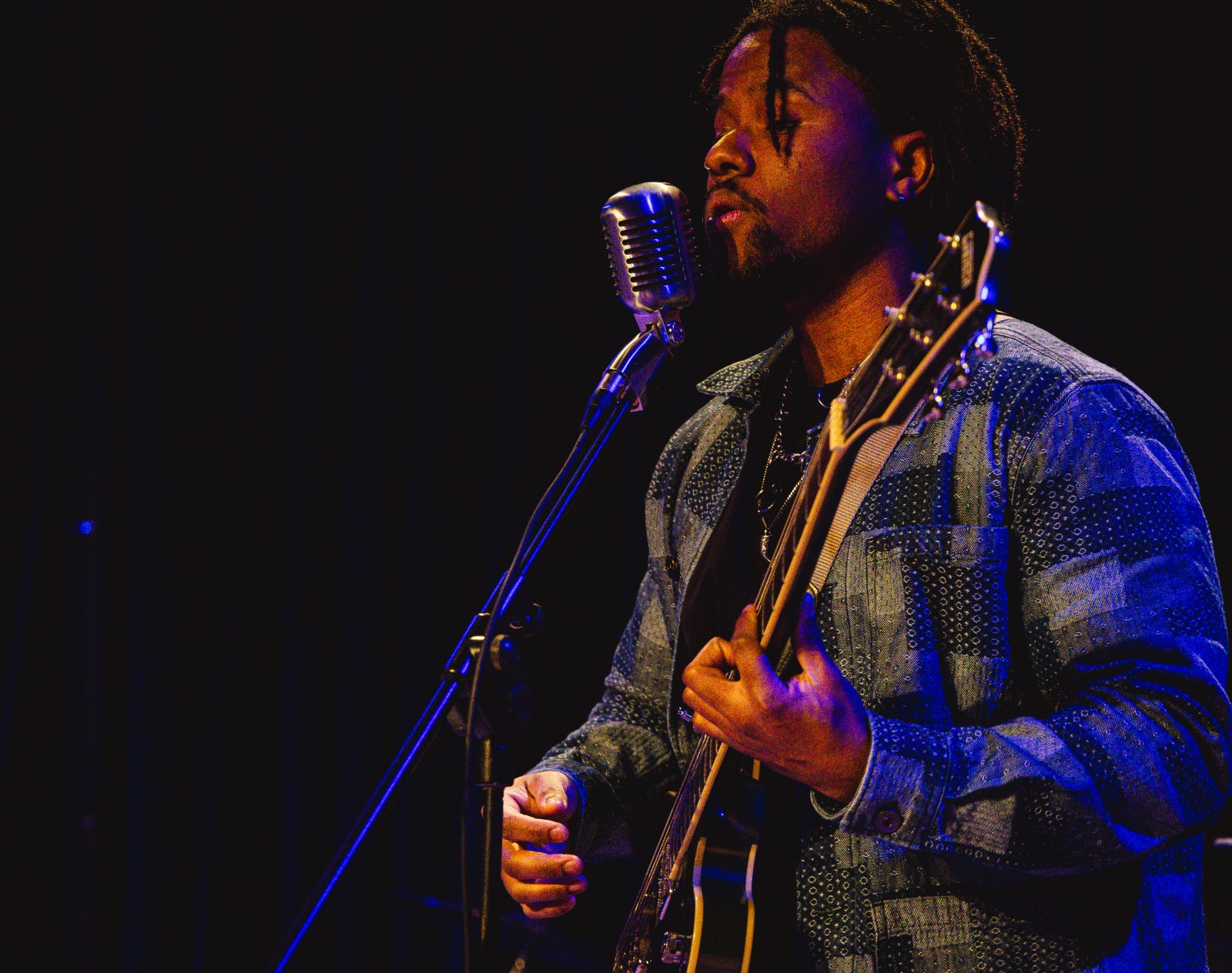 S3BA singing at a vintage microphone with a guitar, in warm stage light.