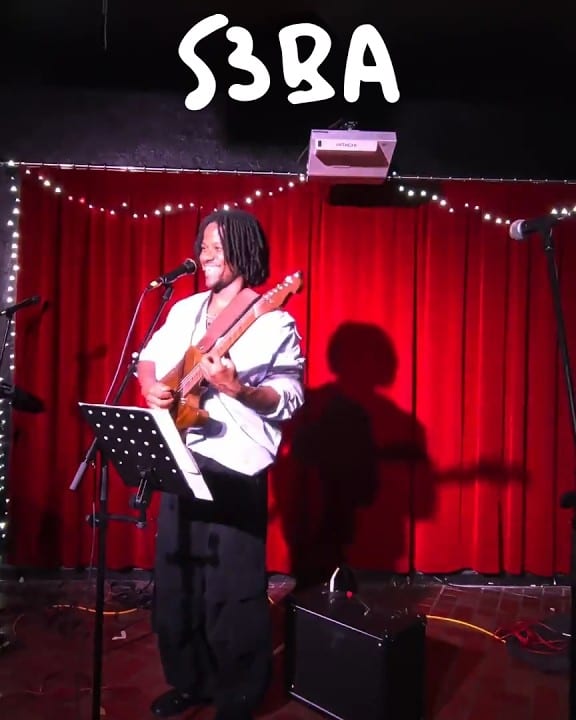 S3BA on stage at Cwps, smiling at the microphone with a guitar in front of a red curtain.