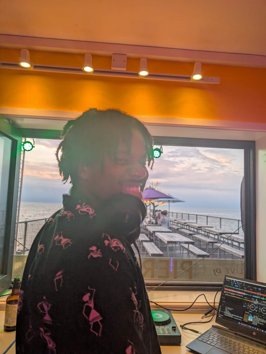 DJ S3BA grinning over one shoulder in the pier booth, headphones round the neck, the sea and a sunset behind.