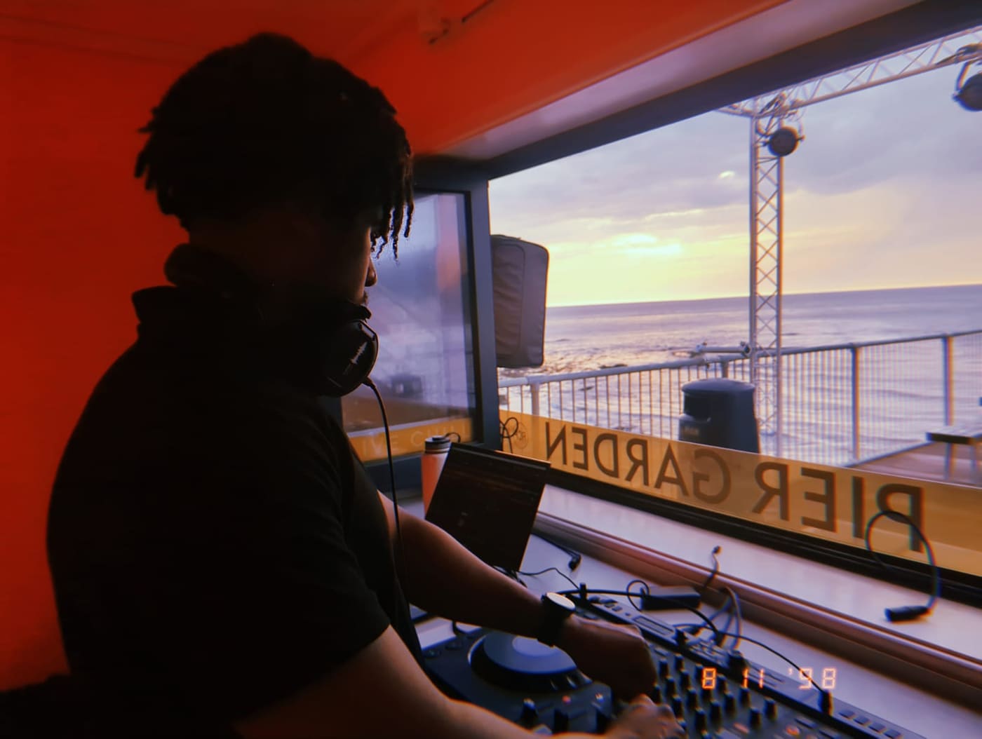 DJ S3BA at the decks in a booth looking out over the sea at sunset.