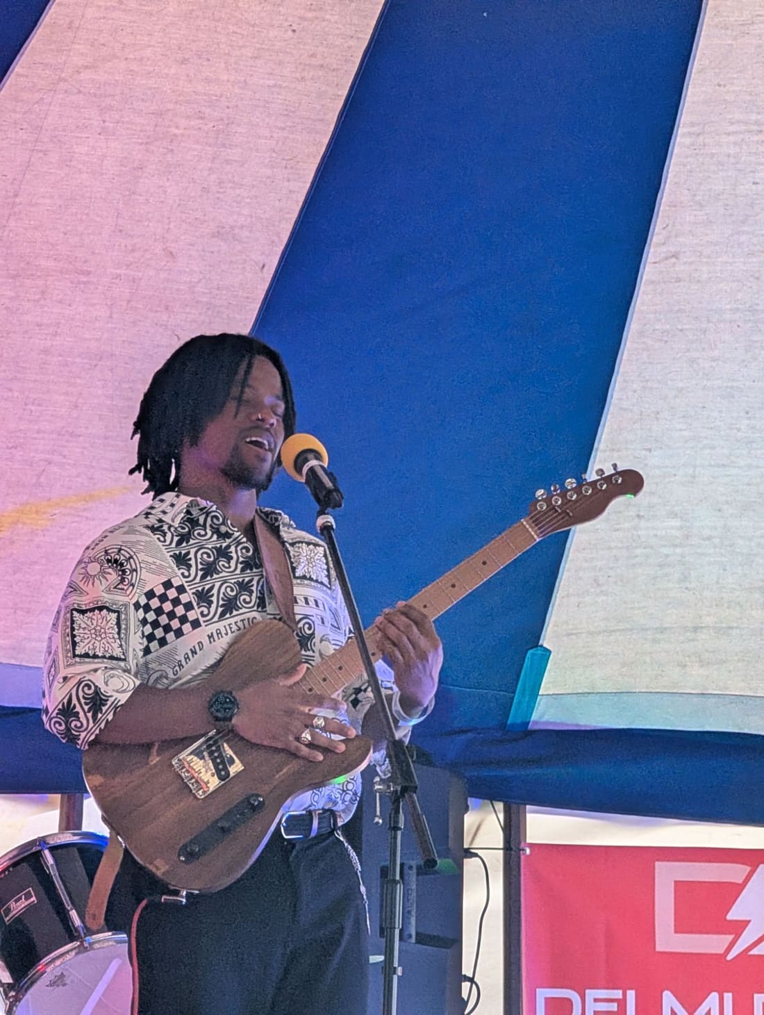 S3BA singing and playing electric guitar on stage under a striped tent.
