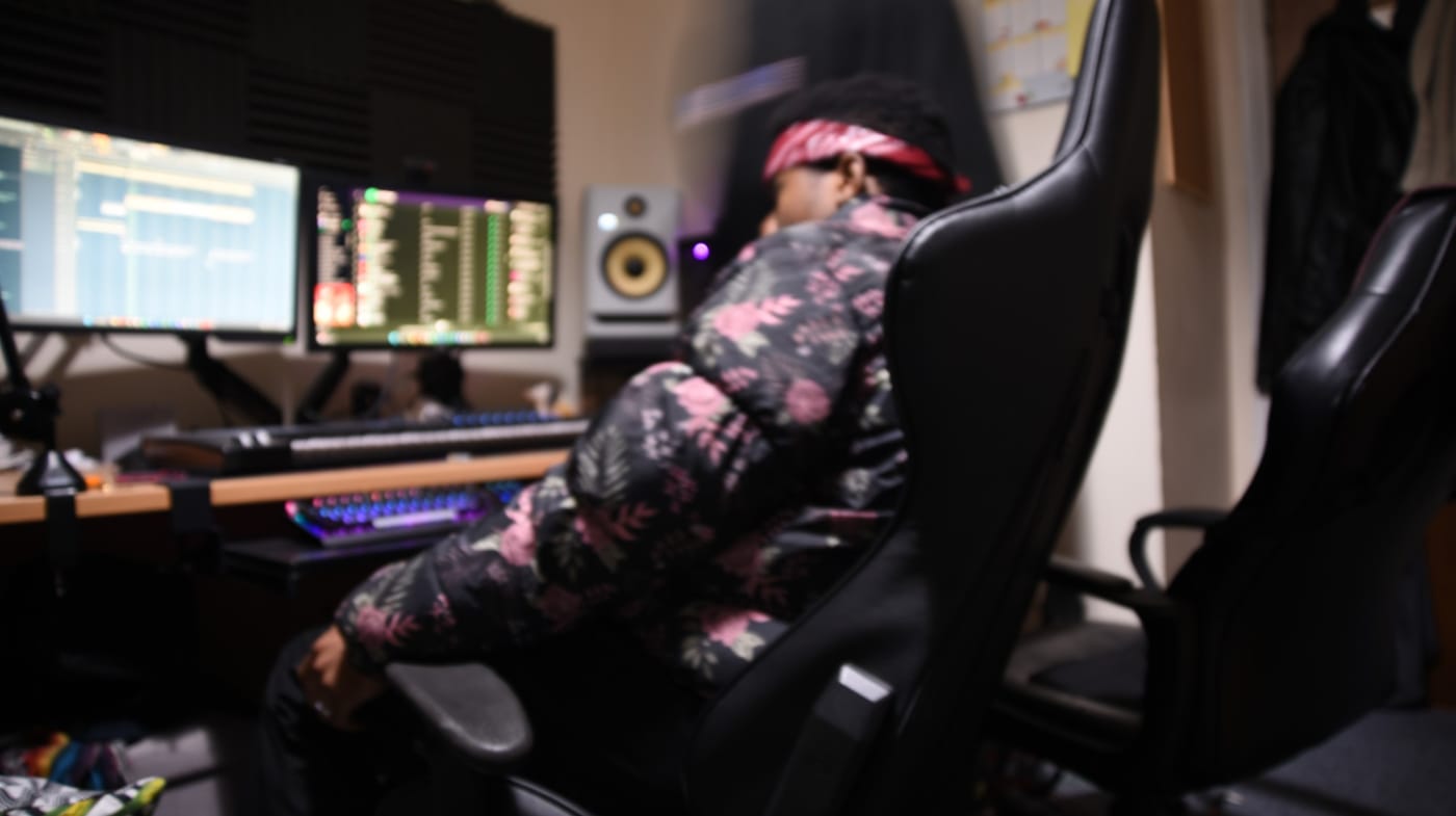 S3BA at the studio desk in a bandana and floral jacket, turned from the monitors, a session open in FL Studio.